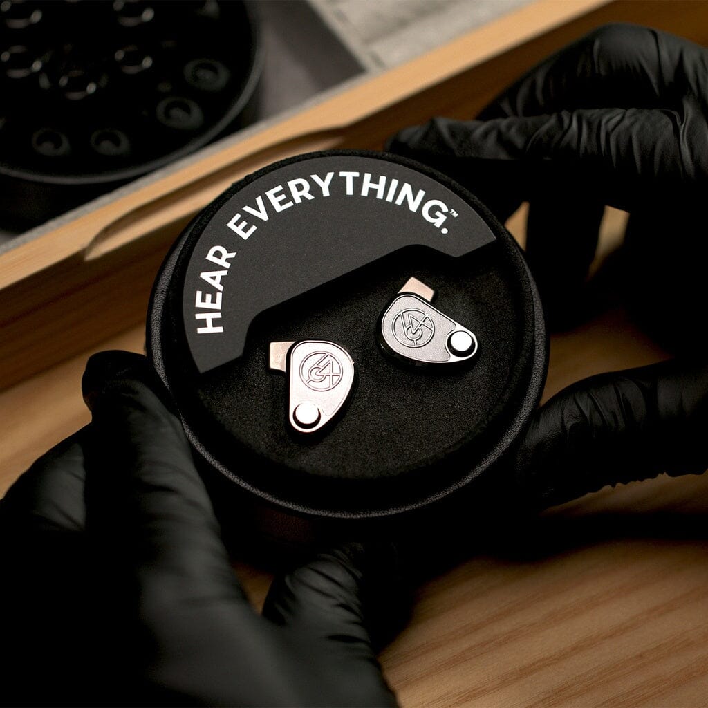 A pair of 64 Audio TWELVE Ti In-Ear Headphones by 64 Audio in a round black HEAR EVERYTHING case, held by gloved hands on a wooden surface.