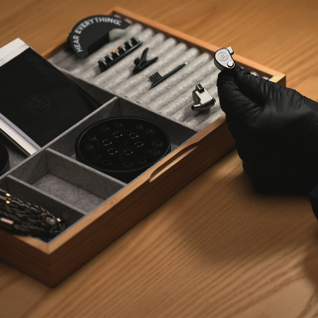 A gloved hand holds a 64 Audio TWELVE Ti In-Ear Headphone above a wooden tray filled with watchmaking tools, parts, and compartments on a light wooden table.