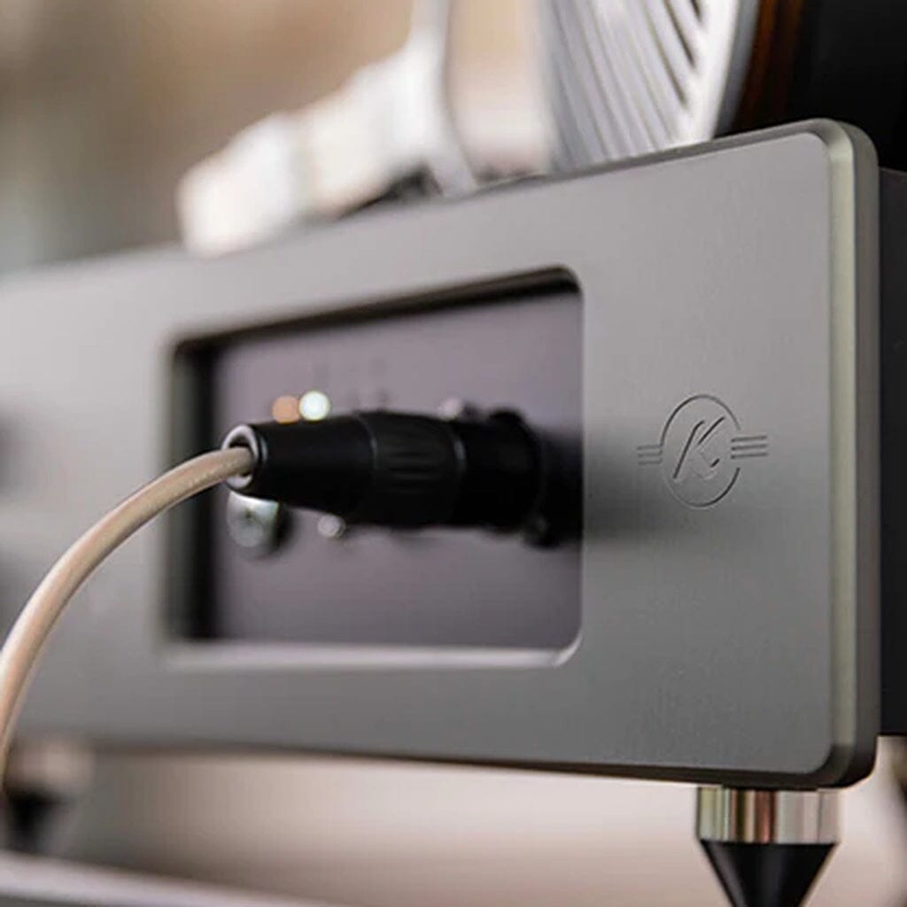 A close-up of the Kallyste Amethyste reference headphone amplifier, an open box clearance item, shows a cable plugged into its sleek chassis and highlights the modern circular Kallyste logo on the side. The contemporary design stands out against a softly blurred background, drawing attention to its advanced features and premium craftsmanship.