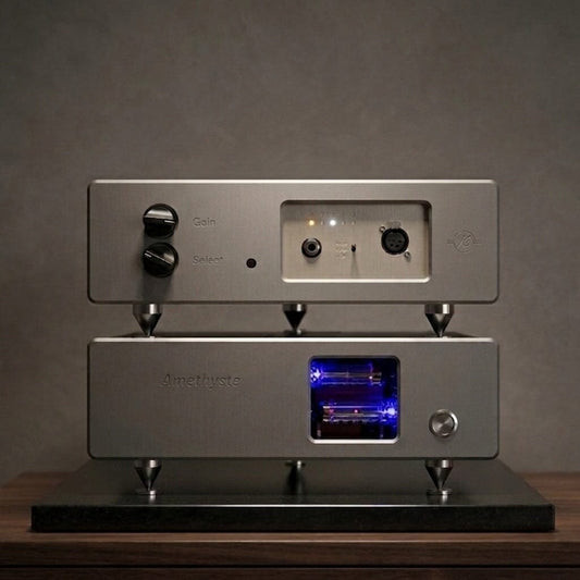 Two stacked silver Kallyste audio components sit on a wooden surface. The top unit, the Kallyste Amethyste—Open Box—Clearance, serves as a reference headphone amplifier with knobs, input ports, and indicator lights; below it is a tube preamplifier featuring a glowing blue-lit window that displays its vacuum tubes.