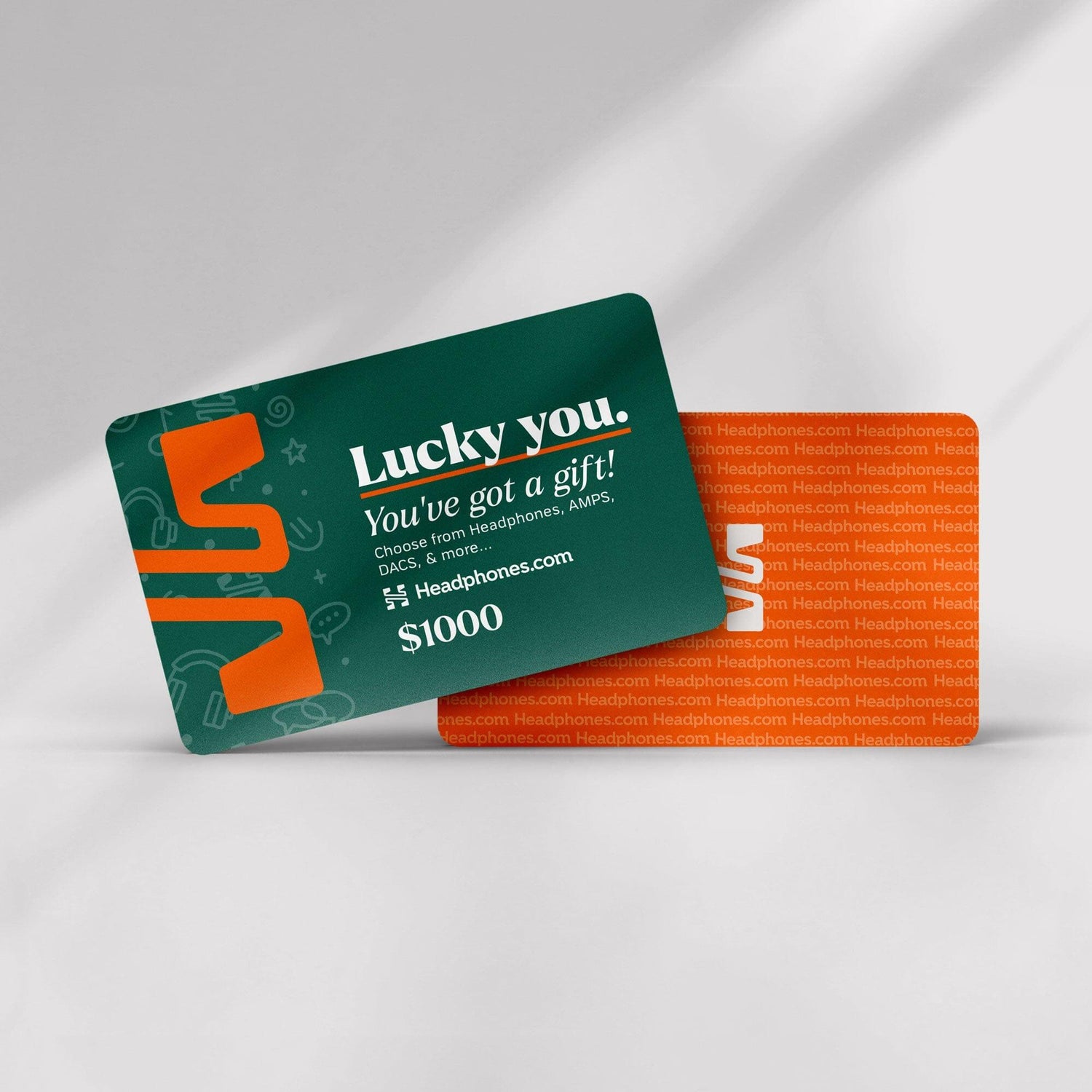 A green headphones.com gift card with “Lucky you. You’ve got a gift!” in white and $1000 displayed is next to an orange Headphones.com card, both resting on a white surface—ready to redeem for your next audio upgrade.